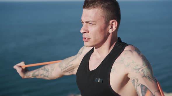 Portrait of Tattooed Man Trains with Elastic Bands at Sunrise with Sea View