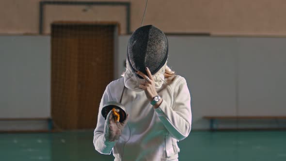 A Young Woman Fencer with Long Blonde Hair Takes Off a Protective Helmet Holding a Sword