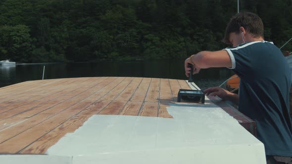 Young man applies white primer on wood boat wheelhouse roof using roller