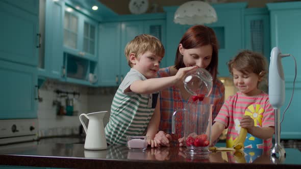 Family of mother and two kids cooking smoothie of strawberries, banana, milk and yogurt with blender