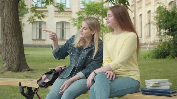 Two Cheerful Caucasian Female Students Sitting in College Yard and Looking Away. Portrait of Young