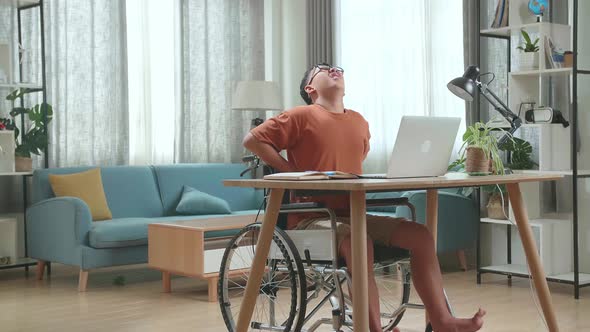 Young Asian Boy Sitting In A Wheelchair Using Laptop Computer And Pain His Back At Home