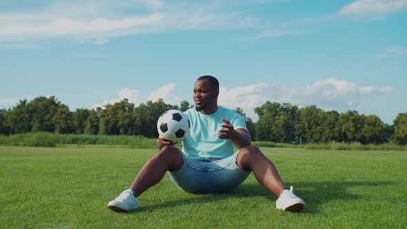 Positive Man with Soccer Ball Sitting on Green Field