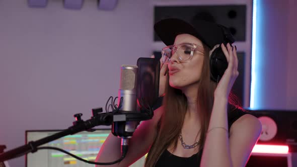 Beautiful Woman in Designer Glasses Cap and Headphones Artist Sings Song in Recording Studio