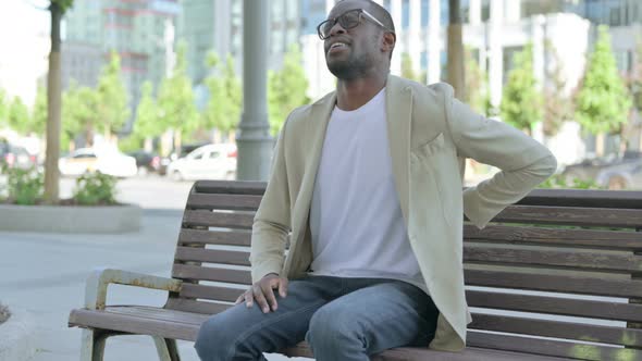 African Man Having Back Pain While Sitting on Bench Outdoor