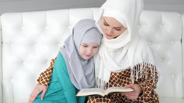 Muslim Mom in Hijab is Reading Her Little Daughter a Book Sitting on the Sofa