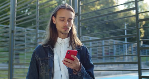 Crop View of Bearded Young Man Using Smartphone While Standing Near Sports Ground. Hipster Guy with