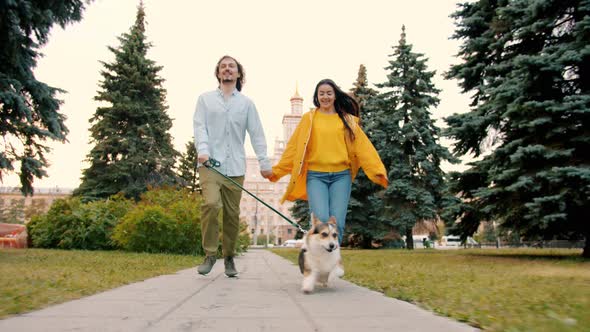 Dolly Shot of Man, Woman and Dog Corgi Breed Running Outdoors in City Park