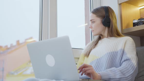 Young woman, at the window of the house, it is snowing. Wearing headphones, working on a laptop.