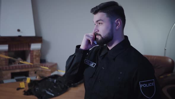 Bearded Caucasian Young Police Officer Standing at the Crime Scene and Talking on the Smartphone
