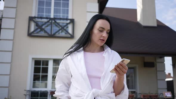 Portrait of Caucasian Woman Smiling Using Smartphone Standing in Front of House for Sale Outdoors