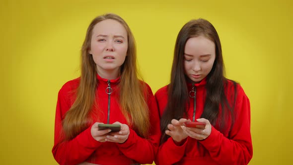 Absorbed Twin Sisters Texting Fast on Smartphones Looking at Camera with Serious Facial Expression