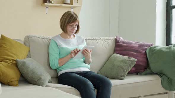 Smiling Elderly Woman Wearing Glasses Using Digital Tablet at Home Sitting on the Couch