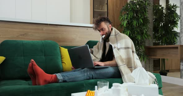Sick Male Freelancer Sitting on Sofa at Home Office Under Plaid Working Remotely Using Paper Tissue