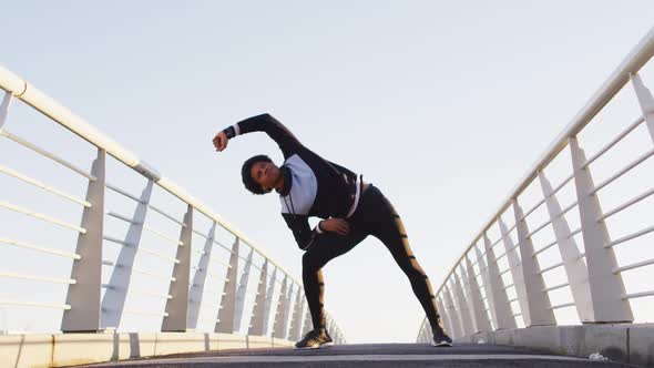 African american man exercising outdoors, standing on footbridge stretching from waist