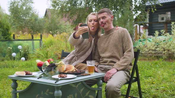 A Young Couple Takes a Selfie at a Picnic Table in the Country on a Smartphone, Shows the Language