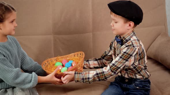 A Blond Caucasian Boy in a Cap Gives a Wicker Basket with Easter Eggs to a Girl