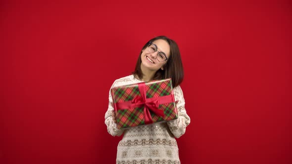 A Young Woman with Glasses Shakes a Christmas Present in Her Hands and Rejoices. Shooting in the