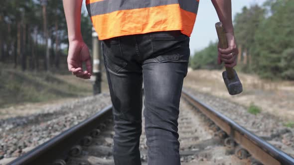 The railway worker walks along the railroad tracks, holding a sledgehammer in his hands.