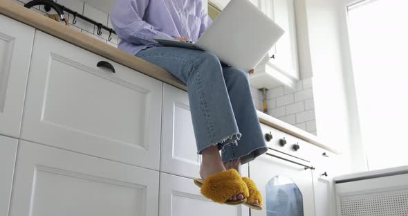 Woman is sitting in kitchen workplace desk and working on laptop drinking coffee.