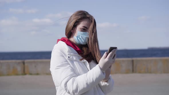 Young Brunette Caucasian Woman in Face Mask and Protective Gloves Using Smartphone. Cute Girl