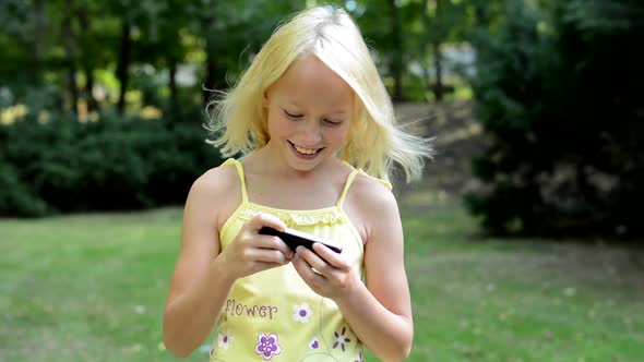 Little Cute Girl Plays the Game on the Smartphone in the Park - She Moves with Phone