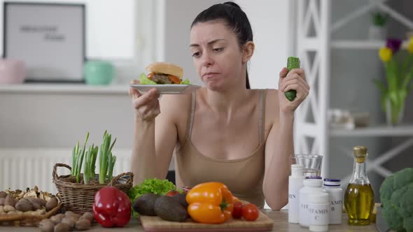 Sad Hungry Young Woman Admiring Hamburger Eating Cucumber Sitting at Table at Home