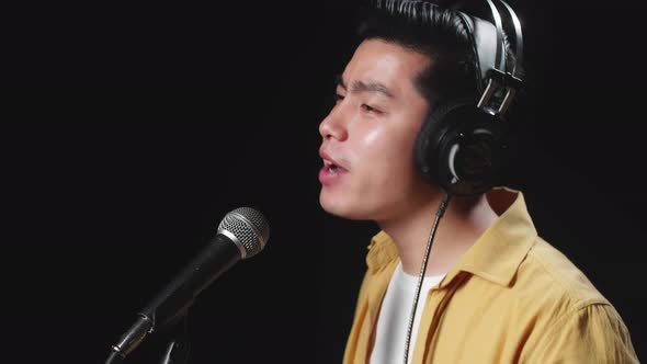 Side View Of Asian Man Singer Wearing Headphone And Singing Into Microphone On Black Background