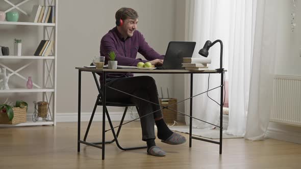 Wide Shot Portrait of Cheerful Laughing Man in Headphones Typing on Laptop Keyboard and Talking