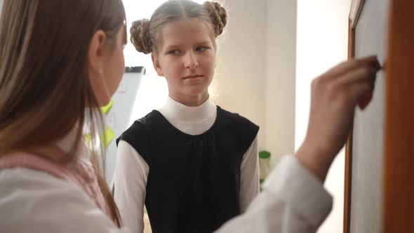 Unsure Girl Shrugging Shoulders As Curios Classmate Talking Writing with Chalk on Blackboard in