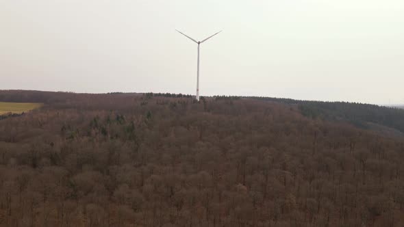 Spinning wind turbine on the top of a ridge behind numerous leafless deciduous trees. Aerial approac