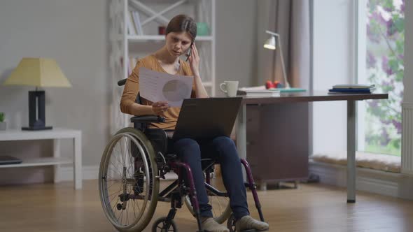 Focused Genius Caucasian Disabled Woman in Wheelchair Talking on Phone Sitting with Laptop and