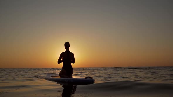 Silhouette of Young Woman Kneeling in Yoga Asana on Supboard in Slowmotion Opposite Sun While Sea is