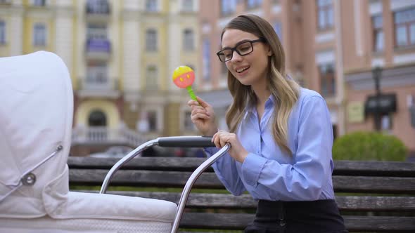 Irritated Working Mother With Rattle Toy Comforting Infant in Pram Sitting Bench