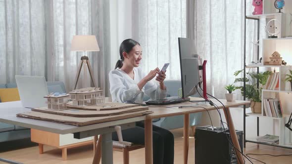 Asian Woman Engineer With The House Model Using Smartphone While Working On A Desktop At Home