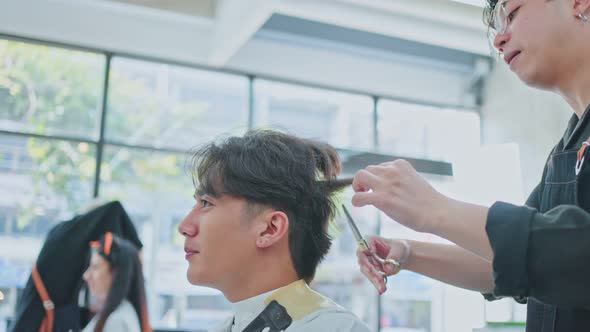 Asian male hairdresser hairstylist combing and using scissors cutting young customer's hair in salon