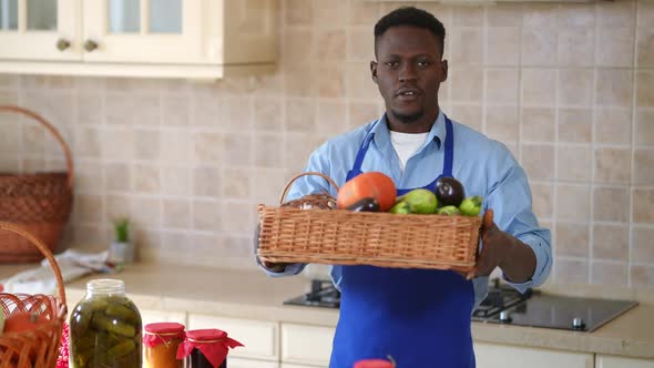 Positive African American Man in Apron Stretching Basket with Pumpkin Eggplant and Garlic Looking at