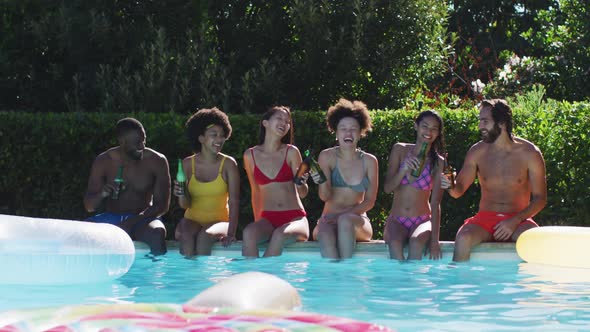 Diverse group of friends sitting at the poolside drinking beer and talking