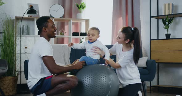 Smiling Caring Mixed Race Parents Having Fun Together with Their Small Kid Putting Her on Fitball