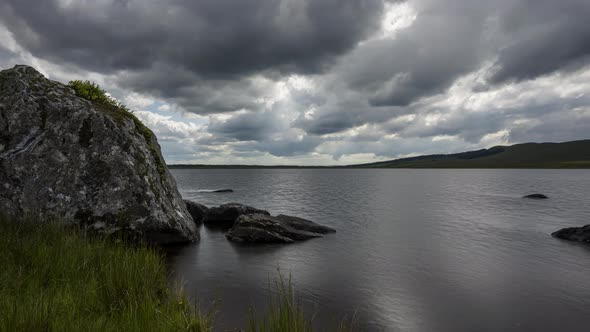 Time lapse of lake with grass and large rocks in the foreground on a dark cloudy summer day in rural alt