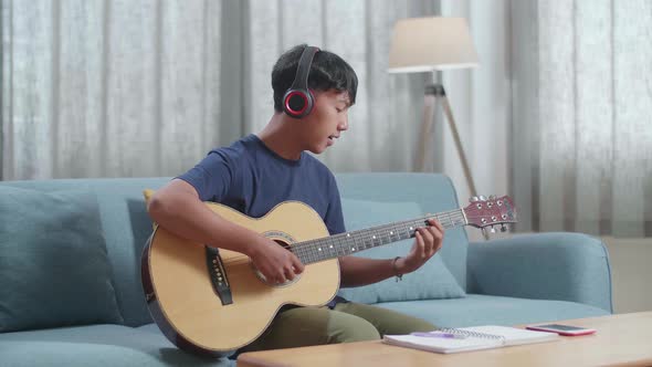 Asian Boy Composer With Notebook On Table Wearing Headphones Singing And Playing Guitar At Home