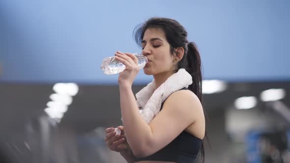 Perspiring Young Fit Middle Eastern Sportswoman Drinking Water Walking in Gym on Treadmill