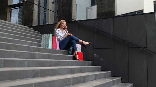 Girl Sitting on Stairs with Bags Talking on Mobile Phone About Sale in Shopping Mall in Black Friday