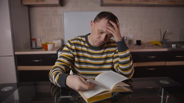 A man in a striped sweater reads a book while sitting at a table, on a blurred background