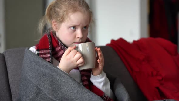 Portrait of a Sick Child in a Scarf and Plaid with a Cup of Hot Tea on the Sofa in the Apartment