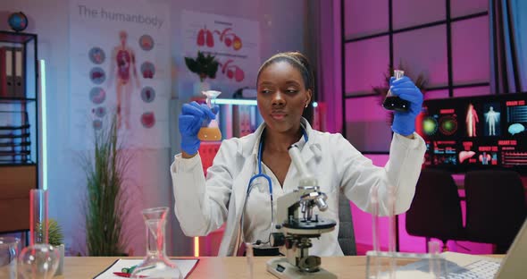 Scientist in Uniform Sitting at the Desk in Lab and Working with Flasks with Chemical Fluids