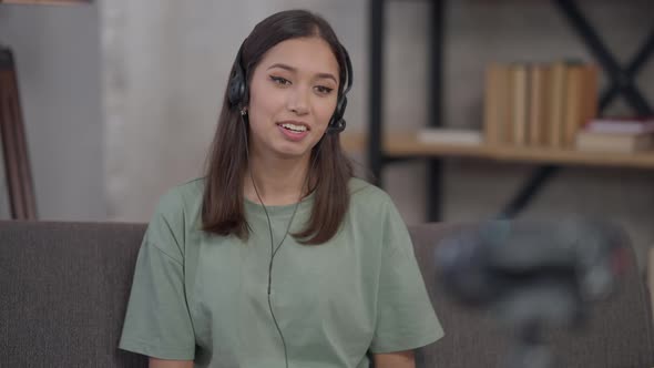 Portrait of Slim Confident Charming Korean Young Woman in Earphones Talking at Video Camera in Home