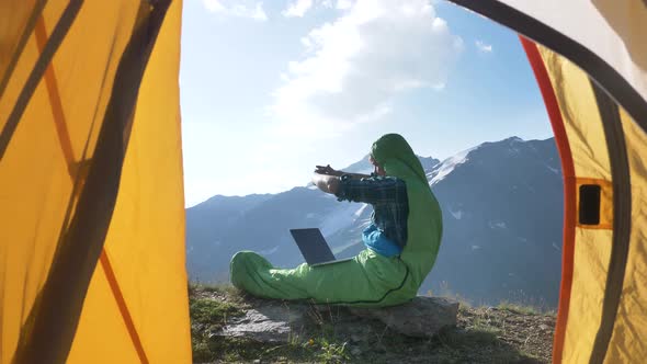 Positive Freelance Man in a Sleeping Bag Opens a Laptop and Starts Working at Dawn in the Mountains