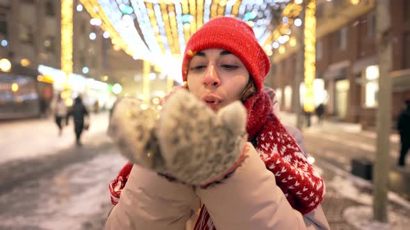 Cheerful Woman Blowing Confetti in Slow Motion on City Square with Festive Illuminations at Winter