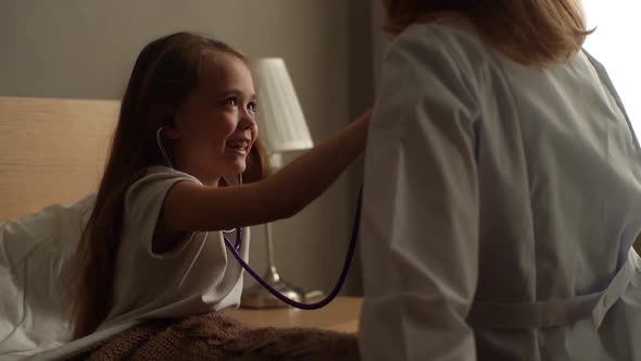 Adorable Cheerful Little Girl Listening Heartbeat with Stethoscope to Female Doctor Sitting at Home